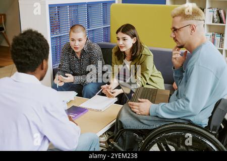 Group of diverse students brainstorming and discussing assignment around study table in library with bookshelves and open laptop, creating inclusive learning environment Stock Photo