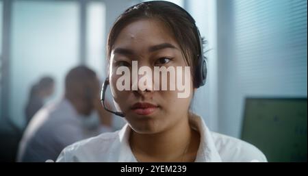 Close up portrait of Asian woman in headset working in call center office. Serious female technical customer support specialist or hotline operator sits at workplace, looks at camera. Client service. Stock Photo