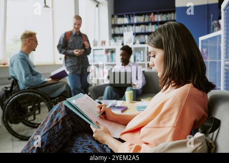 Group of students sitting and studying in a modern, bright library, engaging in conversation and using electronic devices. Multiracial individuals working collaboratively in inclusive setting Stock Photo