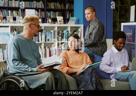 Group of young diverse students collaborating and studying with books and laptops in modern library setting featuring bookshelves and desks. Senior student engaging in conversation Stock Photo
