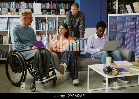 Group of diverse students collaborating in modern library environment discussing projects, sharing ideas seated on sofas and using laptops for studying, learning together Stock Photo