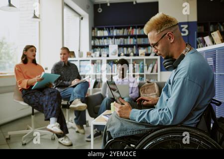 Group of diverse individuals engaging in study session in modern library workspace. Man in wheelchair focusing on work with laptop, while others discussing and taking notes Stock Photo