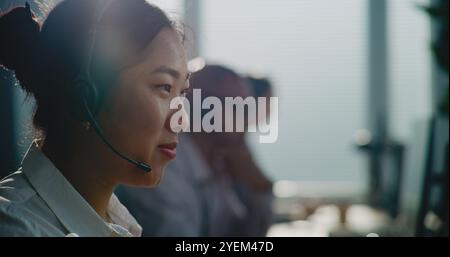 Close up of Asian E-commerce manager in headset having conversation on call with customer, providing online assistance for client. Team of technical support specialists work in call center office. Stock Photo