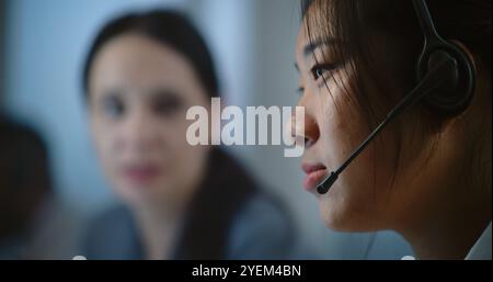 Portrait of friendly female technical support specialist talking to colleague. Asian woman in headset having conversation with customer on call. Diverse hotline operators work in call center office. Stock Photo