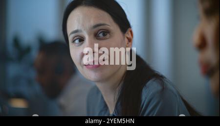 Portrait of friendly female technical support specialist talking to colleague. Asian woman in headset having conversation with customer on call. Diverse hotline operators work in call center office. Stock Photo
