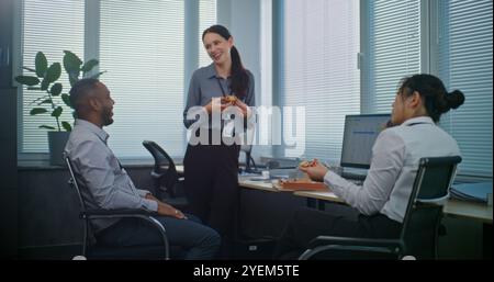 Multiethnic team of technical customer support specialists talk and eat pizza delivery for lunch. Group of company employees resting during break in modern call center office equipped with computers. Stock Photo
