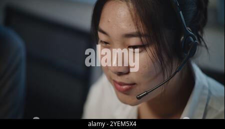 Call center office: Close up portrait of Asian female technical customer support specialist talking on headset, working on computer, providing online assistance for client. Hotline operator working. Stock Photo