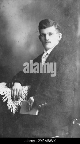 Budapest, Hungary, circa 1905: Elegant portrait of a young man sitting at a table in a photo studio on Mester Street (Mester utca). He is holding a book and sitting in a classic Thonet-style bentwood chair, showcasing the sophisticated urban fashion of the Austro-Hungarian Empire during the Belle Époque. Stock Photo