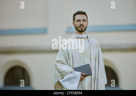 Young Caucasian priest in golden white robe holding Bible smiling lightly while posing for portrait outside Stock Photo