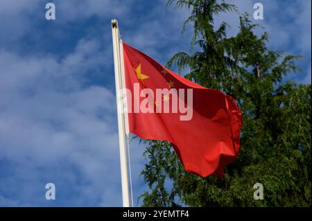 A bright red flag with golden stars flutters in the breeze, set against clear blue skies and lush greenery. Stock Photo