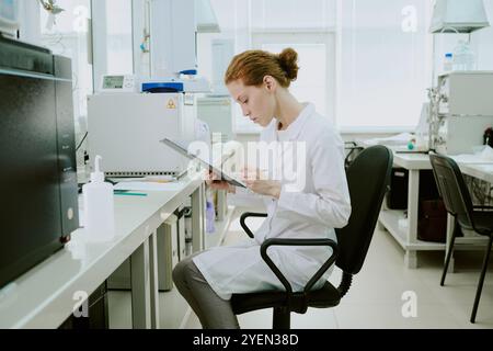 Woman scientist in white coat analyzing data in modern laboratory setting, using a tablet while seated on an office chair, surrounded by scientific equipment Stock Photo
