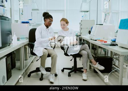 Two scientists sitting in laboratory, analyzing data on tablets while discussing research. Laboratory equipment and samples placed on counters around them Stock Photo