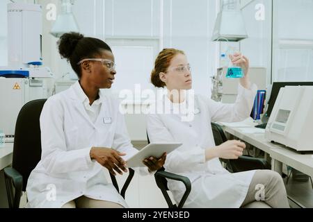 Two female scientists working on scientific research in a laboratory environment, analyzing a blue liquid solution and checking data on a tablet device Stock Photo
