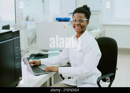 Smiling scientist facing laptop while working in laboratory, wearing protective gear. Equipment and devices in background illustrating research environment Stock Photo