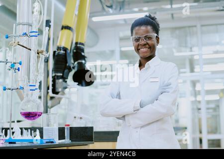 Portrait of Black scientist wearing protective gear while conducting experiment in laboratory workplace. Smiling and standing with folded arms amidst laboratory equipment Stock Photo