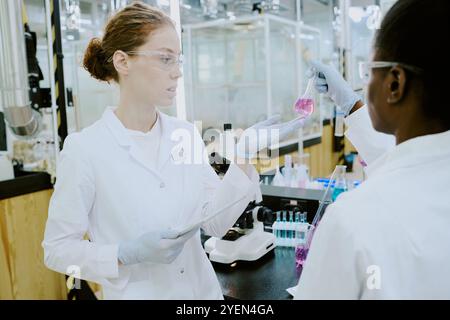 Scientist wearing lab coat examining chemical reaction in beaker while another person observing findings. Laboratory equipment on table with bright lighting creating a professional environment Stock Photo