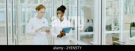 Two female scientists examining documents and tablet while discussing research findings in modern laboratory setting Stock Photo