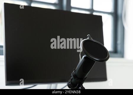 Microphone with pop filter in front of blank computer monitor at home. Podcast, audio, recording, workspace, home studio, broadcast Stock Photo