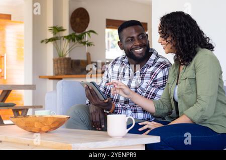 Smiling multiracial couple using tablet together at home, enjoying coffee and conversation. technology, lifestyle Stock Photo