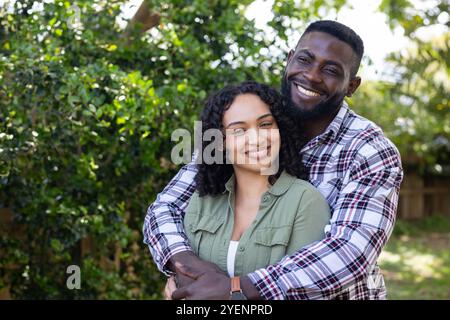 Happy multiracial couple embracing outdoors, smiling warmly in lush green garden. Romance, happiness, love, nature, affection Stock Photo