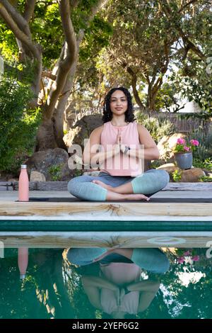 Woman meditating by pool, practicing yoga outdoors with water bottle nearby. meditation, relaxation, wellness, mindfulness, exercise, fitness Stock Photo