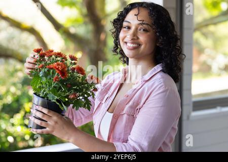 Smiling woman holding potted flowers on porch, enjoying sunny day. Gardening, lifestyle, happiness, outdoors, leisure, relaxation Stock Photo