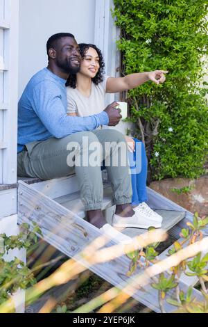 Smiling multiracial couple enjoying morning coffee on porch, pointing at nature. happiness, togetherness, outdoors, relaxation Stock Photo