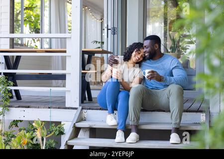 Happy multiracial couple enjoying coffee together on porch, embracing and smiling warmly. love, togetherness, happiness, relaxation, affection, relati Stock Photo