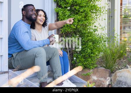 Smiling multiracial couple enjoying coffee on porch, pointing at something outside. morning, relaxation, bonding, togetherness, conversation Stock Photo