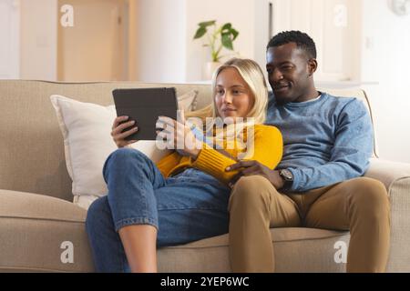 multiracial couple relaxing on couch, watching tablet together at home, feeling cozy. Lifestyle, technology, leisure, comfort, bonding, togetherness Stock Photo