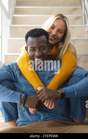 Smiling multiracial couple sitting on stairs, embracing warmly in cozy sweaters, at home. love, romance, relationship, affection, warmth, knitwear Stock Photo