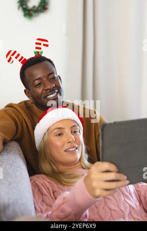 Christmas time, multiracial couple wearing festive hats taking selfie with tablet at home. Holiday, celebration, technology, happiness, self-portrait, Stock Photo