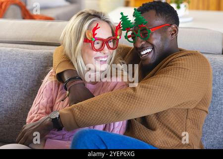 Christmas time, smiling multiracial couple wearing festive glasses, at home. celebration, winter, together Stock Photo