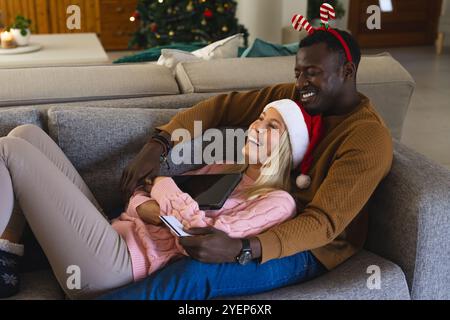 Christmas time, smiling multiracial couple on couch shopping with tablet and credit card, at home. technology, lifestyle, ecommerce, celebrations Stock Photo