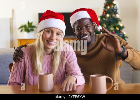 Smiling multiracial couple in Santa hats enjoying holiday coffee at home by Christmas tree. Celebration, winter, festive, cozy, family, sipping Stock Photo