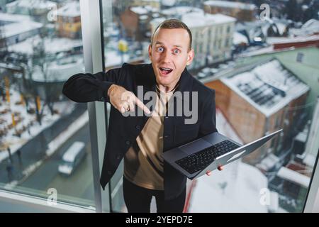 Portrait of a young joyful man with a laptop in his hands, standing near the window. Work in the office. Successful male manager Stock Photo