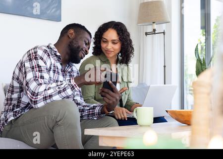 Smiling multiracial couple using smartphone and laptop at home, enjoying cozy time together. Technology, leisure, lifestyle, communication, bonding Stock Photo