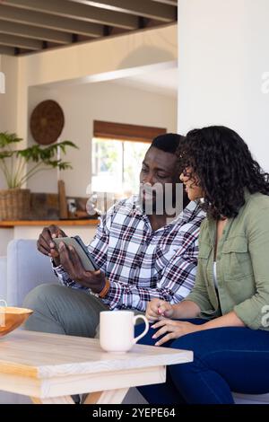 Happy multiracial couple using tablet at home, enjoying coffee and conversation together. technology, communication, relaxation, lifestyle, domestic, Stock Photo