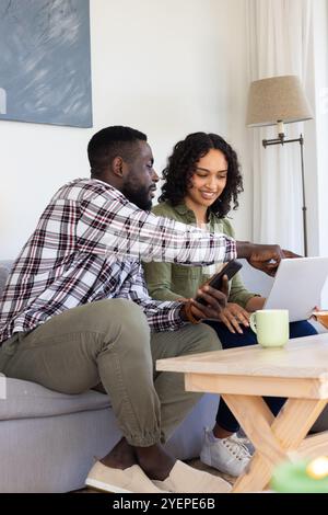 multiracial couple using laptop and smartphone at home, smiling and discussing together. Technology, digital, communication, lifestyle, teamwork, rela Stock Photo