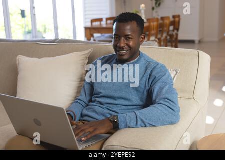 Smiling African american man using laptop on couch at home, enjoying relaxed afternoon. Technology, leisure, relaxation, remote work, lifestyle, casua Stock Photo