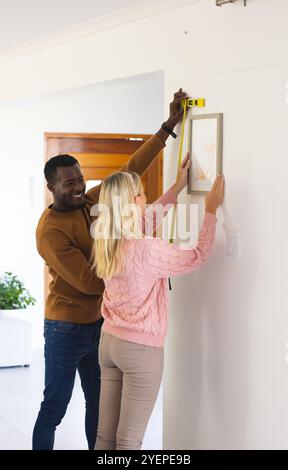 multiracial couple hanging picture on wall, measuring and smiling at home. home improvement, teamwork, interior, lifestyle, decoration, teamwork Stock Photo