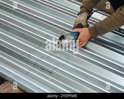 A man in work clothes and a glove uses an angle grinder to cut or carve a metal sheet or roofing material along markings Stock Photo