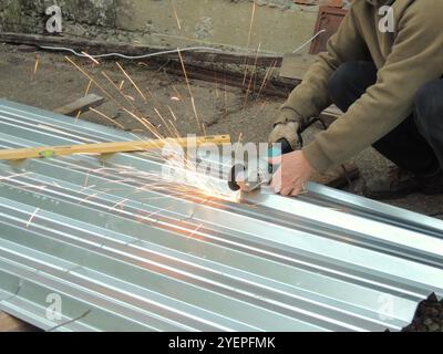 A man in work clothes and gloves uses a grinder to cut or saw a metal sheet or canvas on asphalt, causing sparks to fly Stock Photo