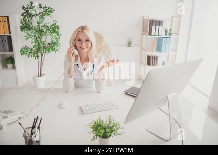 Smiling senior doctor in modern clinic office communicating with patient over phone, offering medical consultation and care Stock Photo