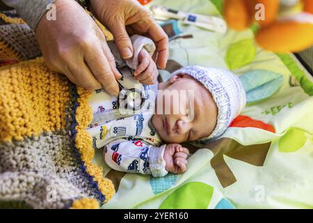 A beautiful baby boy is seen in the first days of life, wrapped in soft blankets during a medical checkup, midwife uses stethoscope listening to chest Stock Photo