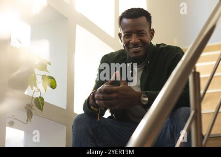 Smiling African american man sitting on stairs at home, enjoying sunlight and relaxation. happiness, casual, lifestyle, tranquility, leisure, indoors Stock Photo