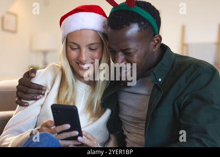 Christmas time, Smiling multiracial couple wearing festive hats looking at smartphone at home. Celebration, technology, lifestyle, happiness, joyful, Stock Photo