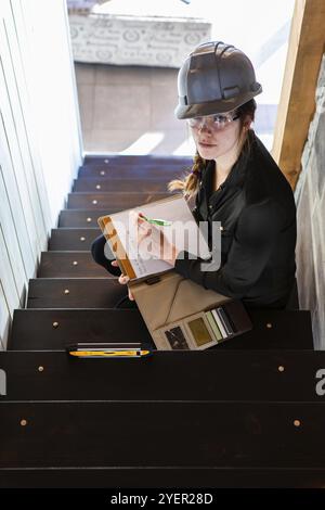 Construction inspector woman sitting on the stair and taking notes on her clipboard. during pre purchase inspection of house interior stairs. vertical Stock Photo