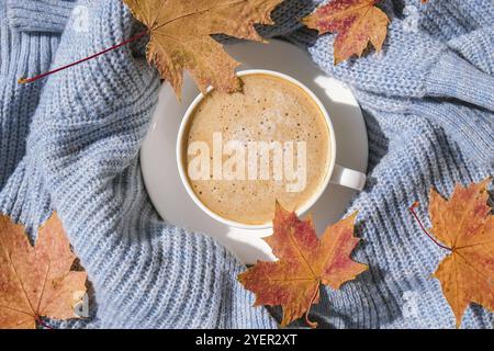 White cup of morning warming coffee on blue knitted sweater with maple yellow leaves background. Cozy home concept. Aesthetics blog lifestyle. Autumn Stock Photo