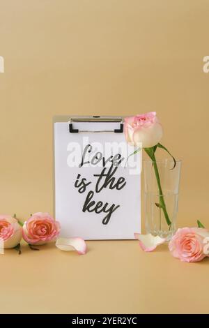 LOVE IS THE KEY text on greeting card paper note. Delicate pink roses on beige background. Minimal trendy composition. Romantic pastel pink rose flowe Stock Photo